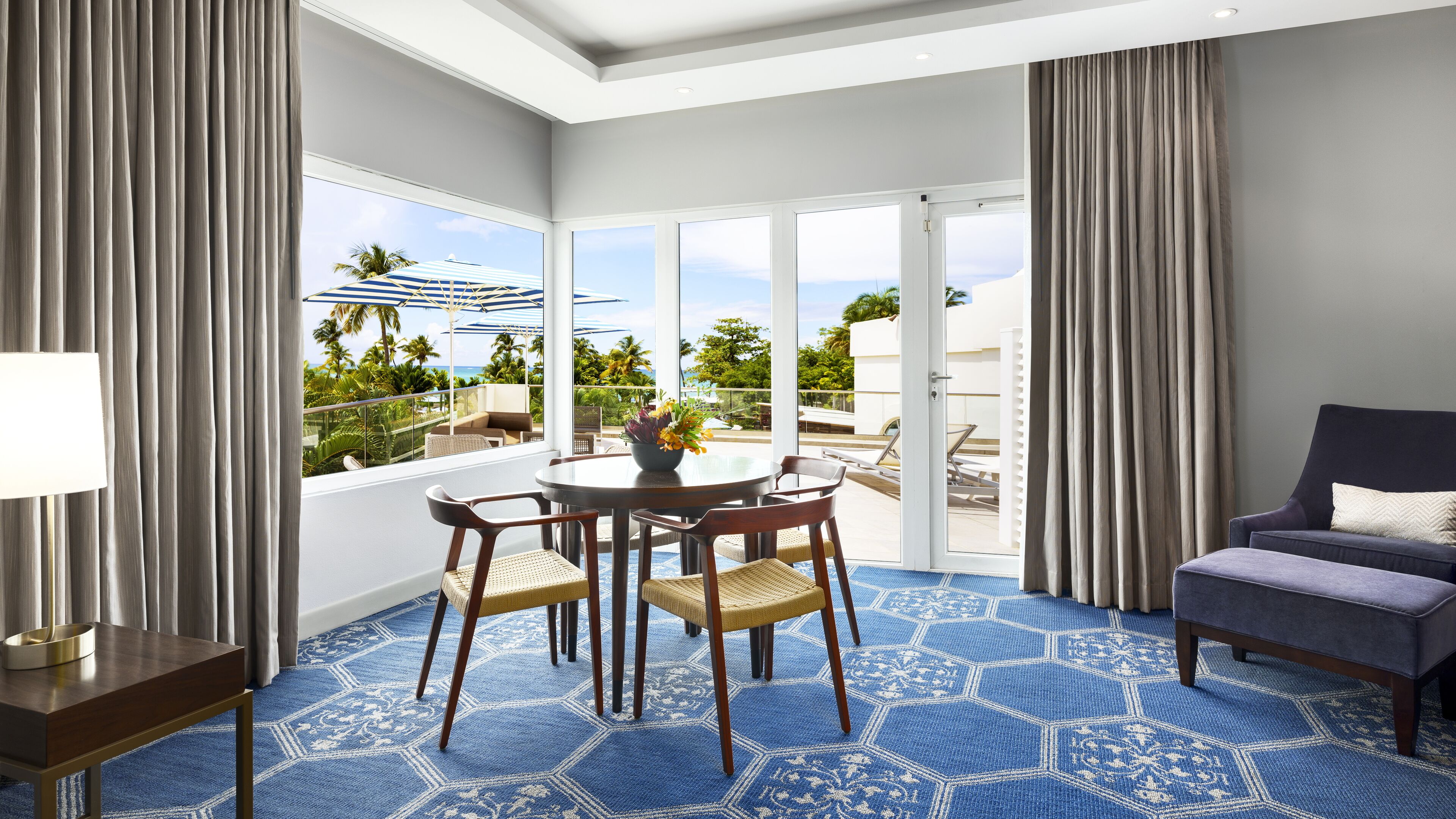 A hotel room with a balcony, a round table with chairs, a sofa, and a view of the ocean and palm trees.