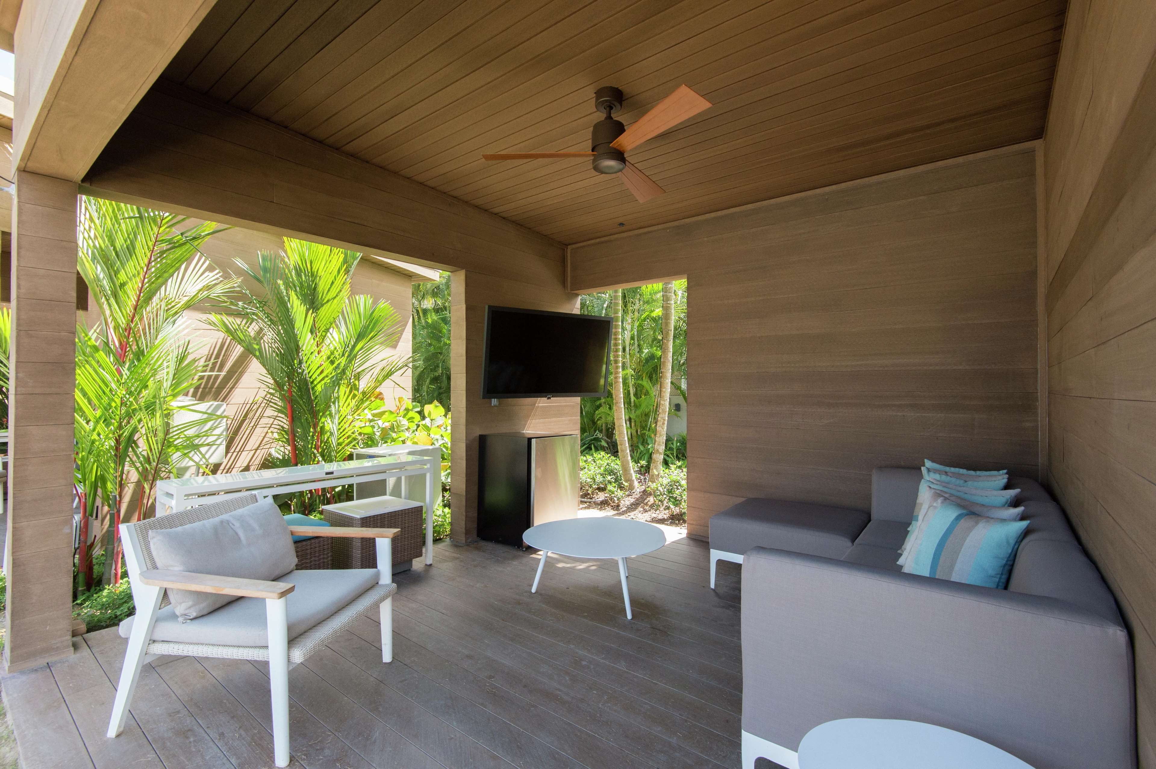 A covered outdoor seating area with a ceiling fan, a flat-screen TV, and a sofa with cushions.
