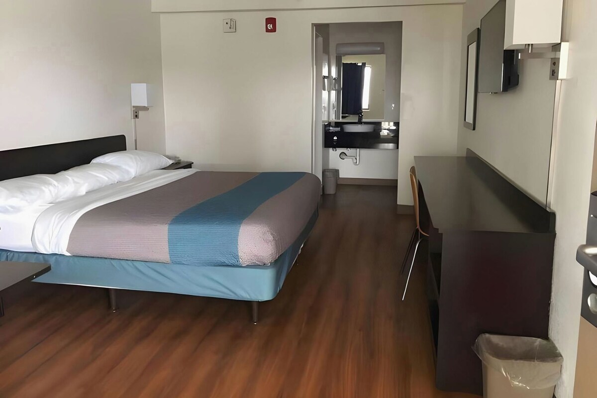 A hotel room with a bed, a desk, a chair, a small table, a trash bin, a mirror, and a bathroom with a sink and mirror.
