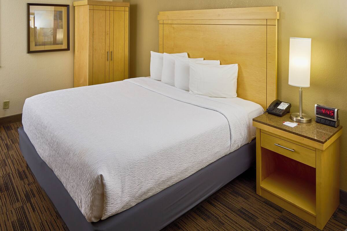 A hotel room with a large bed, wooden headboard, a bedside table with a lamp, a phone, and a digital clock.