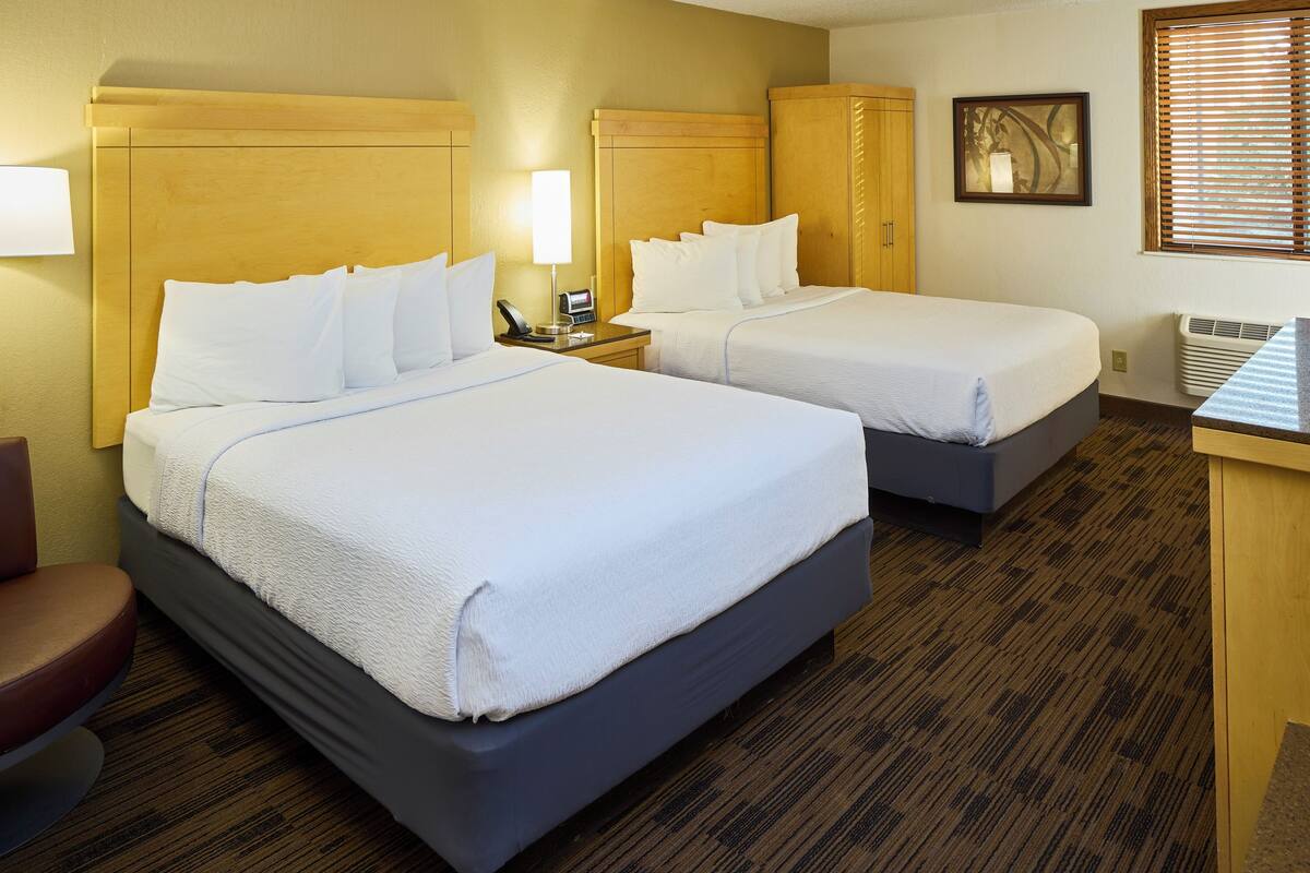 A hotel room with two beds, a desk, a chair, and a window with blinds.