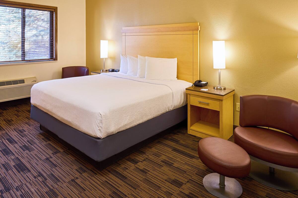 A hotel room with a large bed, bedside lamps, a wooden nightstand, a chair, and a heater.