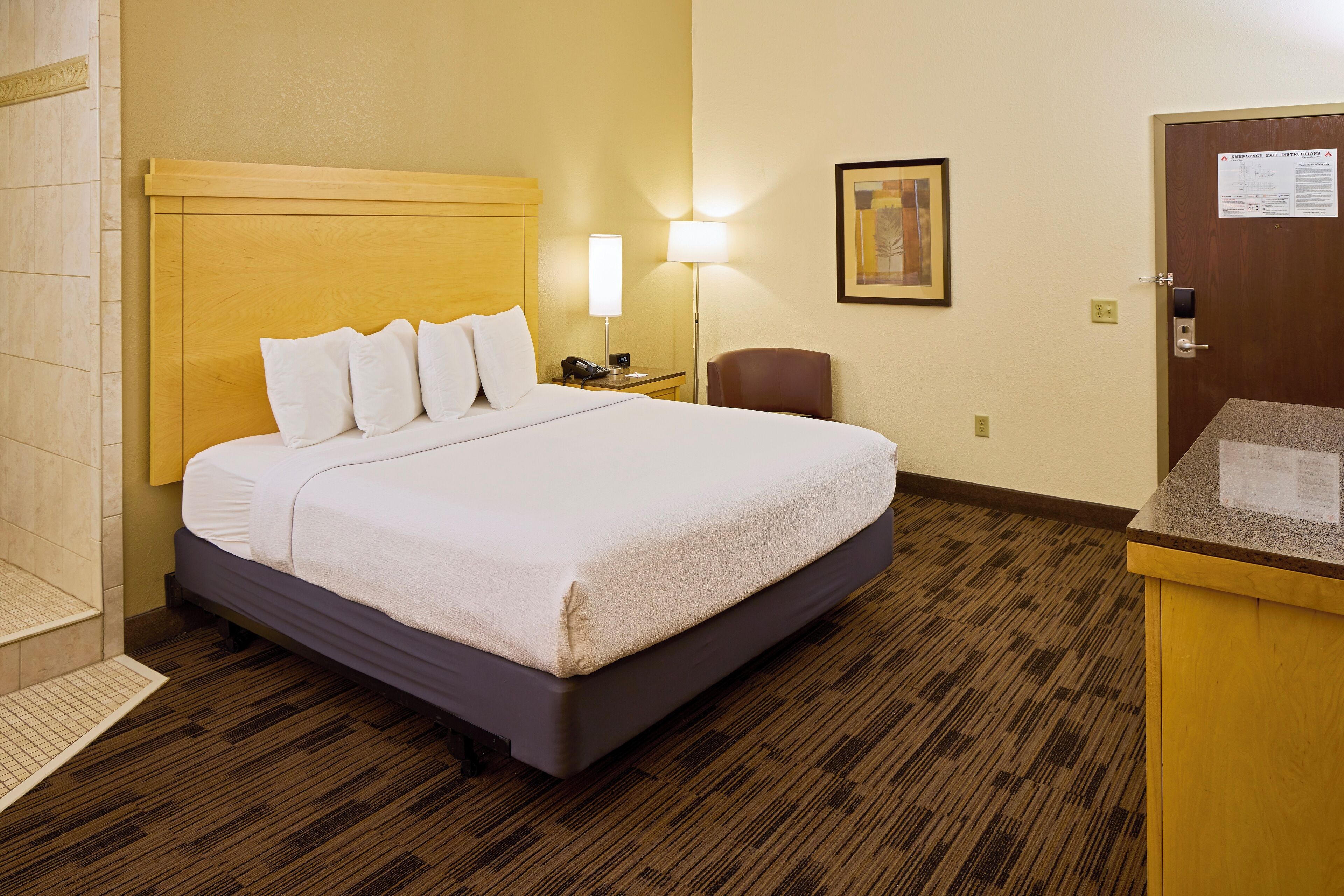 A hotel room with a bed, bedside lamp, chair, and a desk with a computer.