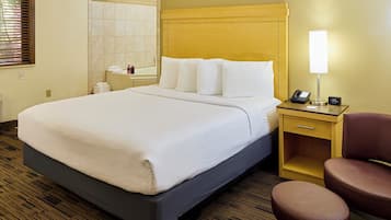 Quarto de hotel com uma cama grande, uma mesinha de cabeceira com abajur, uma cadeira e um banheiro visível através de uma porta em arco.