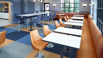 A modern cafeteria with long tables, blue and orange chairs, and a patterned carpet.