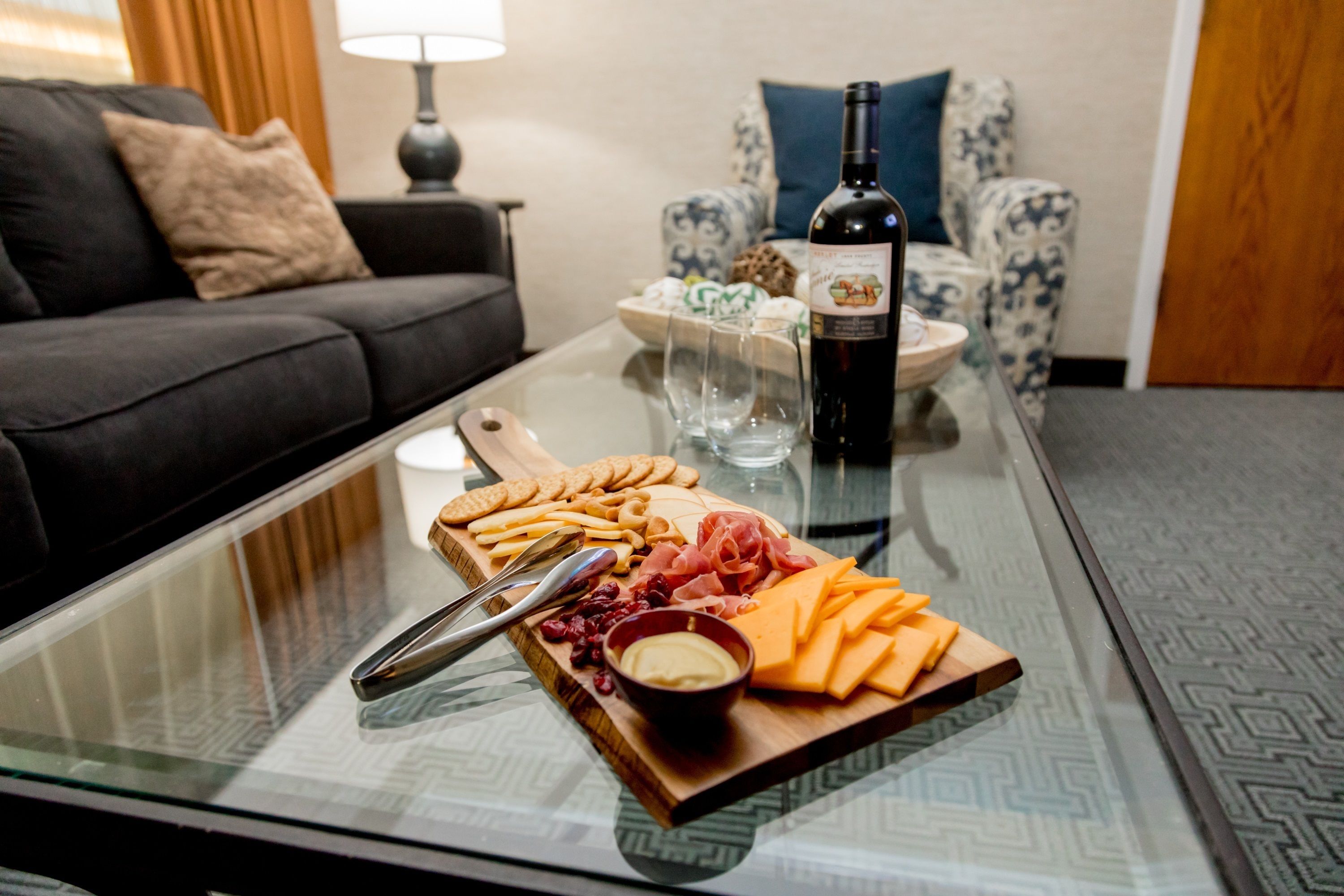 A glass-top table with a charcuterie board, a bottle of wine, and a bowl of dip.