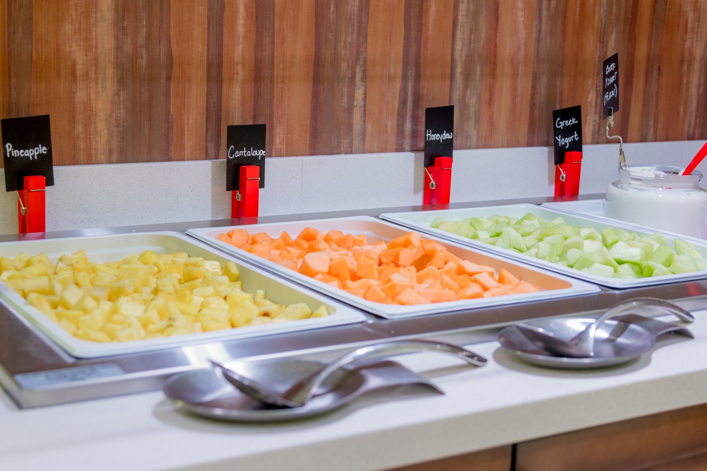Un bufé con recipientes identificados de piña, melón cantalupo, melón honeydew y yogur griego.