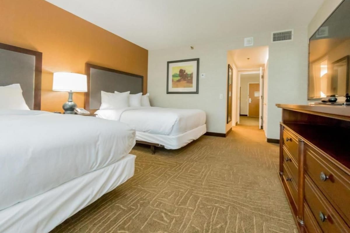 A hotel room with two beds, a dresser, a lamp, a painting, and a door leading to another room.