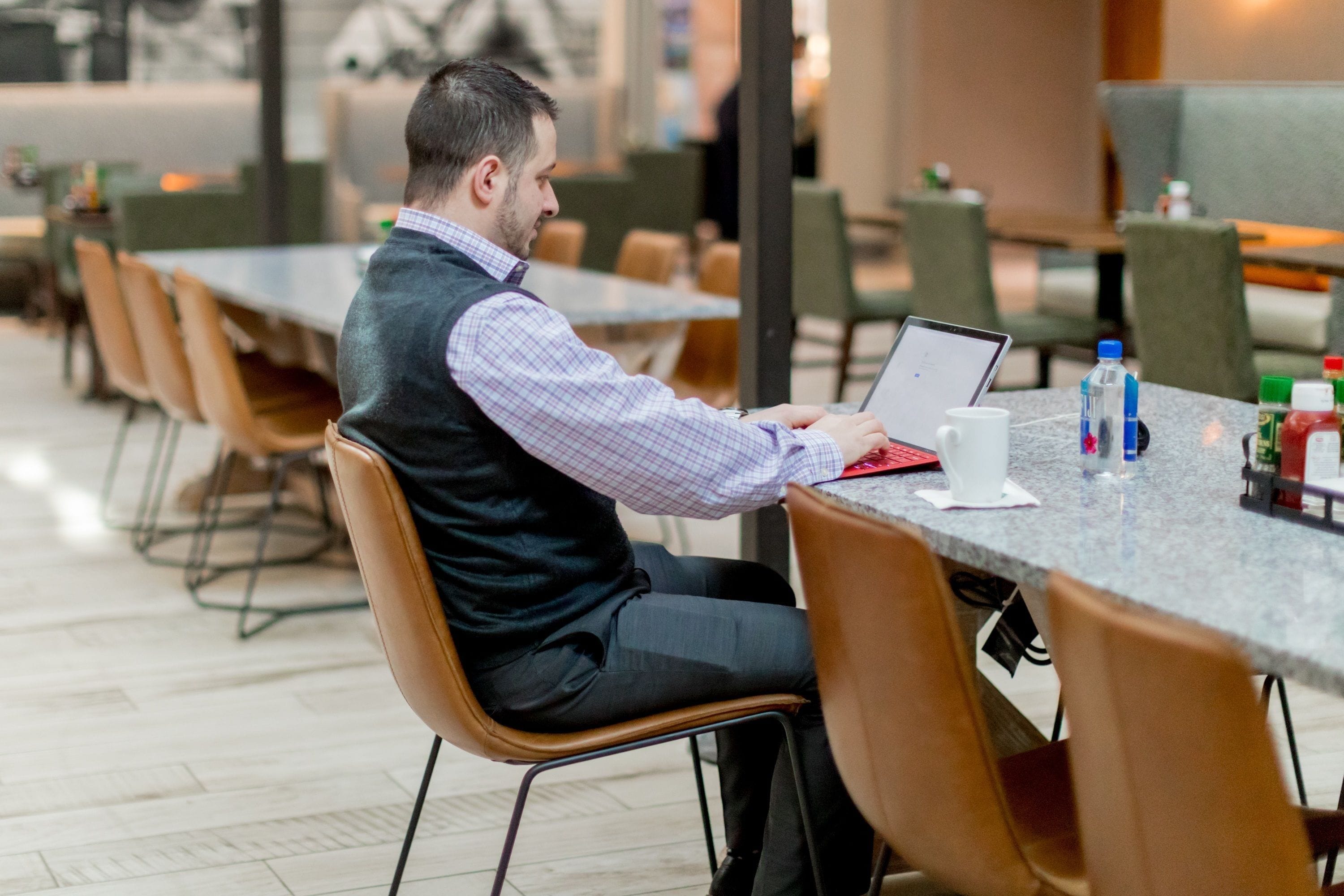 A man seated at a table with a laptop, a notebook, and a cup. The setting appears to be a modern indoor space with chairs and tables arranged in rows.