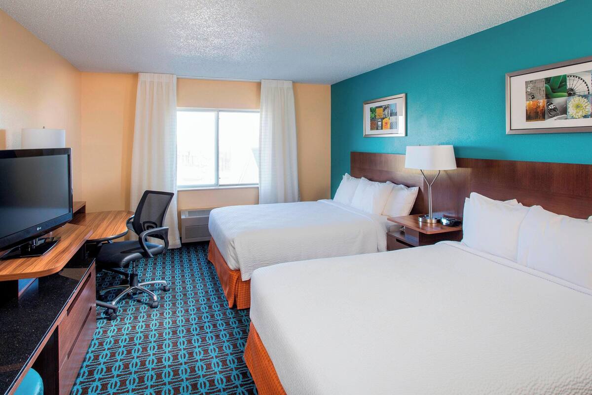 A hotel room with two beds, a desk with a TV, a chair, a window with curtains, and a painting on the wall.