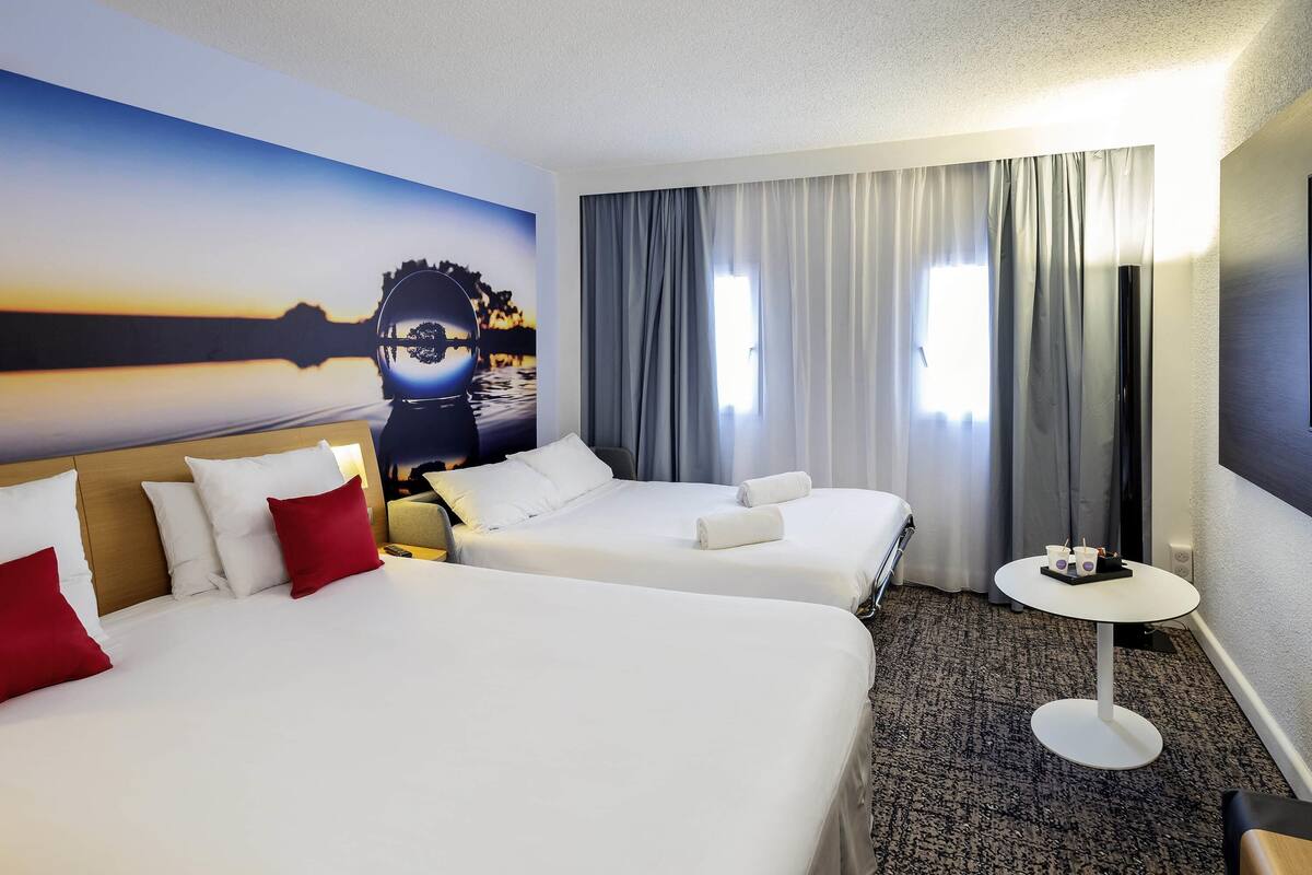 A hotel room with two beds, a small round table, a painting on the wall, and a view of the outdoors through the window.
