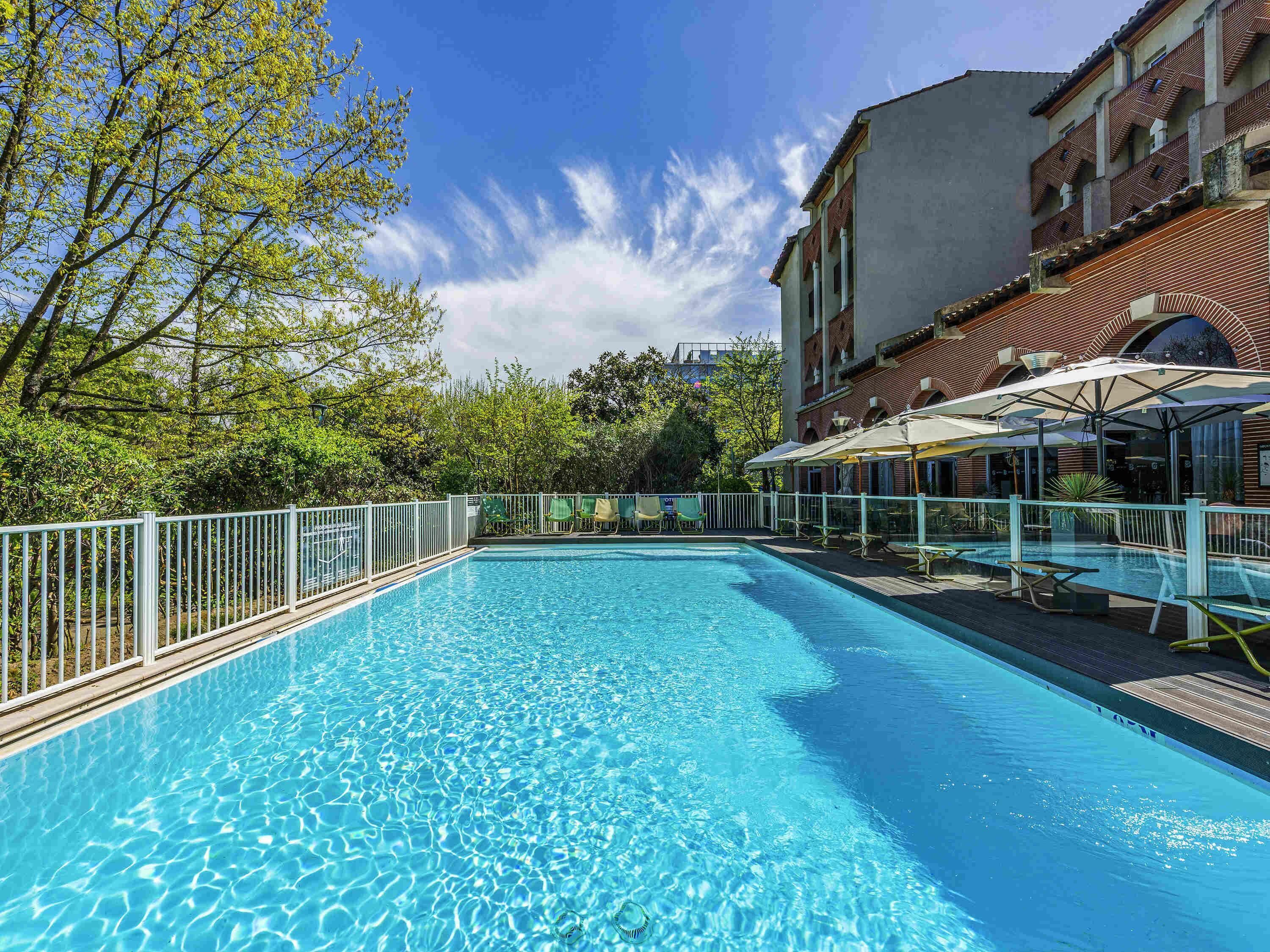 Una piscina azul cristalina con una cerca blanca, rodeada de árboles y un edificio con techo de tejas.