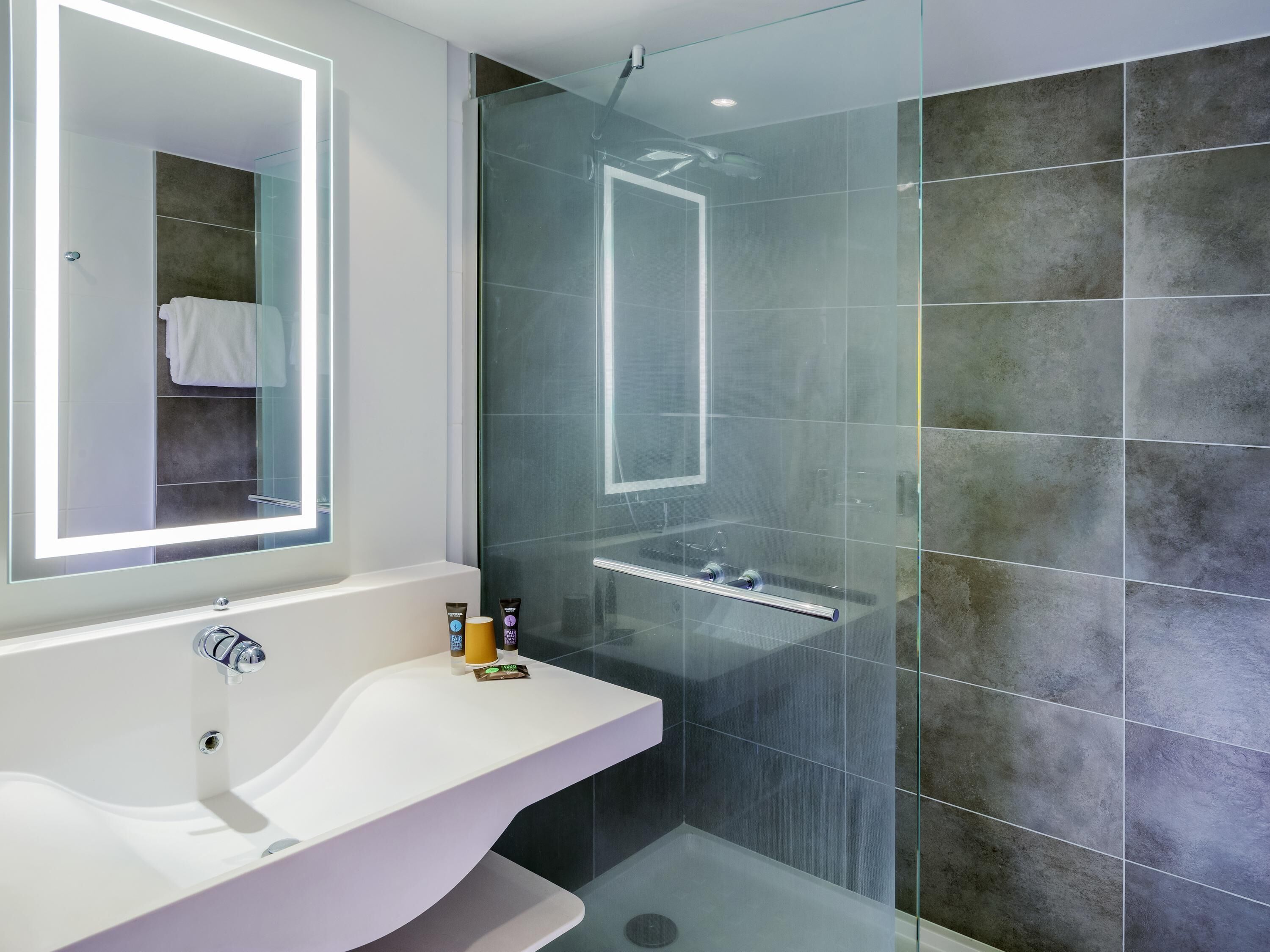 Un baño moderno con ducha envuelta en vidrio, tocador blanco con espejo y pared de azulejos.