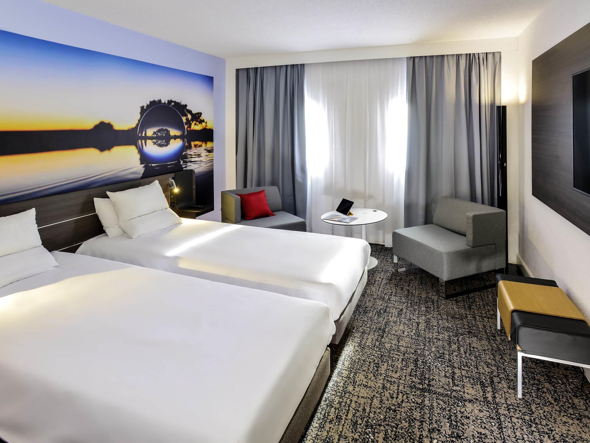 A hotel room with two beds, a sofa, a small table, and a large window with curtains.