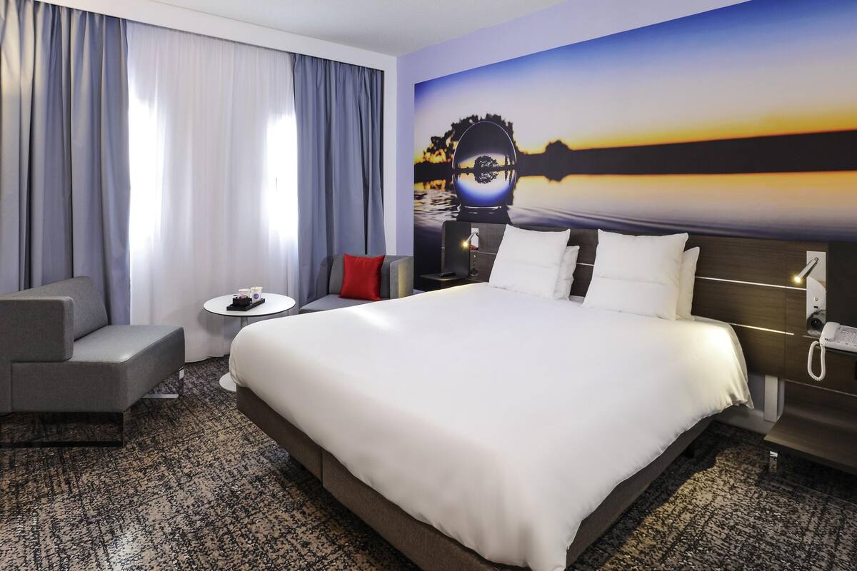 A hotel room with a large bed, a sofa, a small table, and a painting of a sunset.