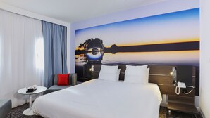 A hotel room with a large bed, a small table, a chair, a nightstand with a phone, and a wall with a scenic painting. - Novotel Toulouse Centre Compans Caffarelli (Toulouse)