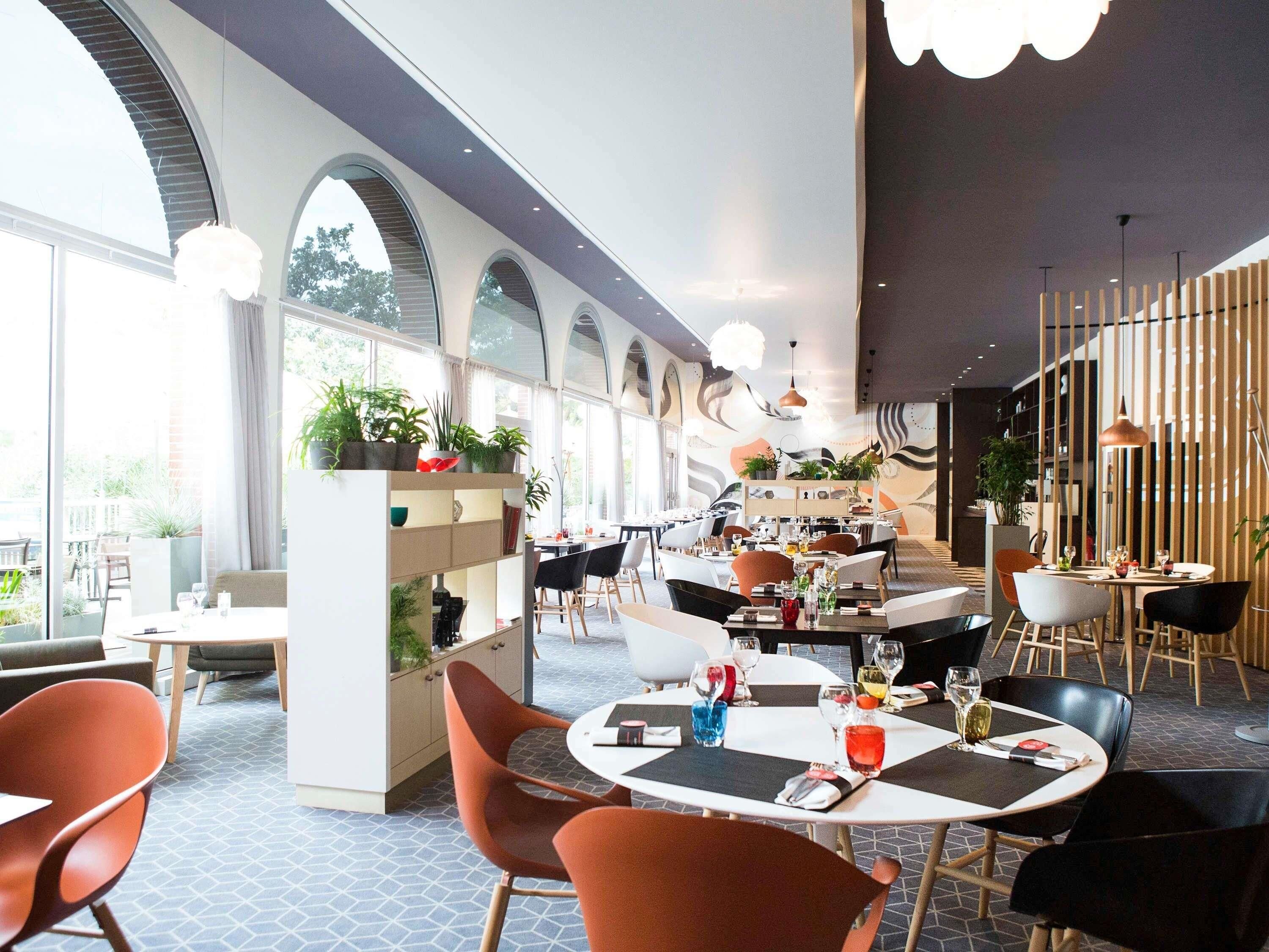 A modern restaurant with high arched windows, indoor plants, and a variety of seating arrangements.