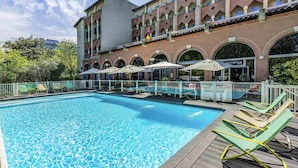 A swimming pool with lounge chairs, a restaurant, and a building with arched windows. - Novotel Toulouse Centre Compans Caffarelli (Toulouse)