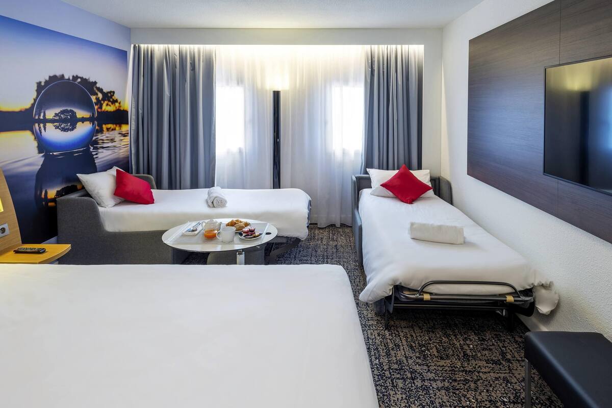 A hotel room with two beds, a small table with breakfast, a TV, and a painting on the wall.