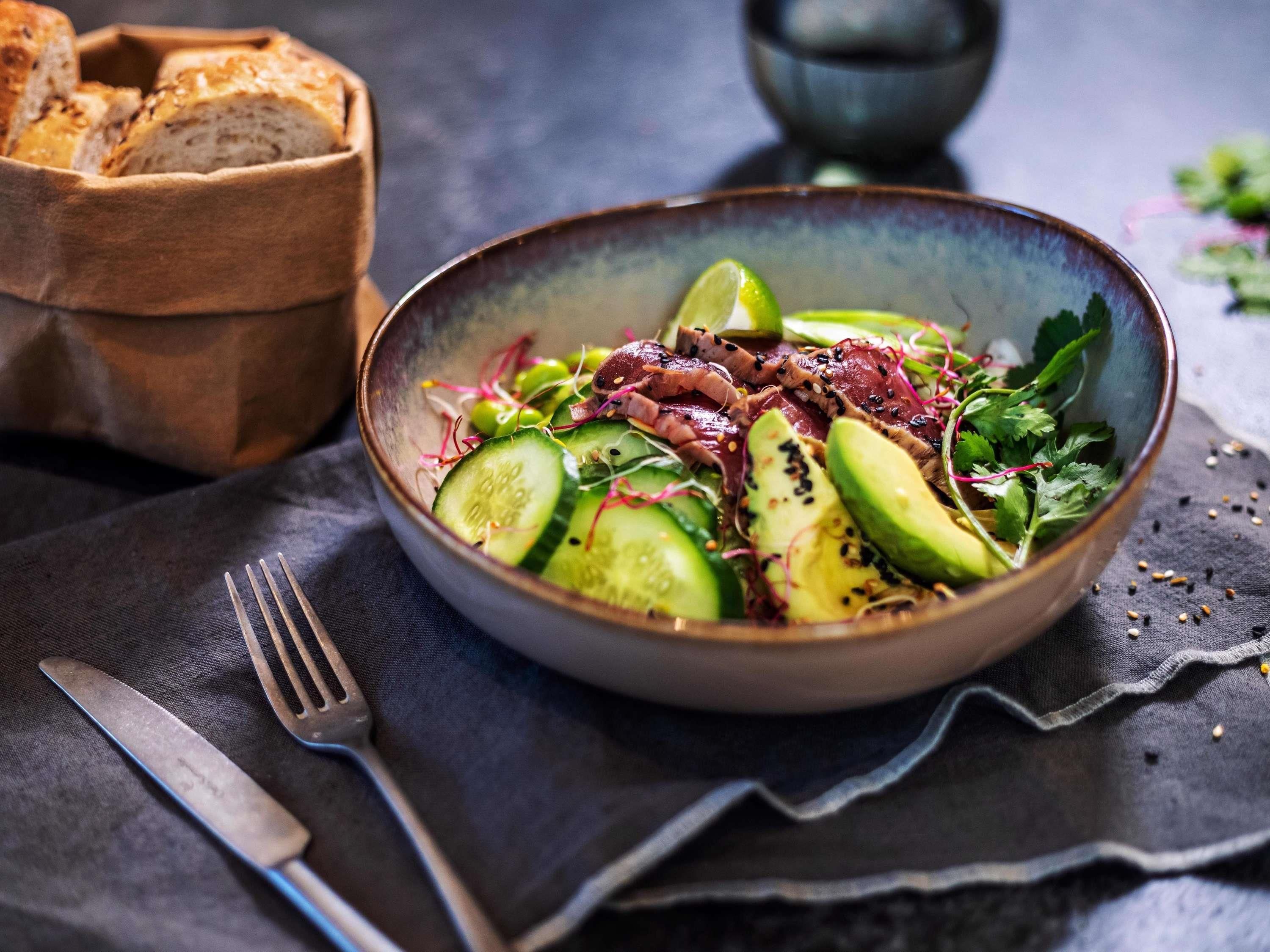 A bowl of salad with various vegetables and meat, accompanied by bread and a knife on a dark cloth.