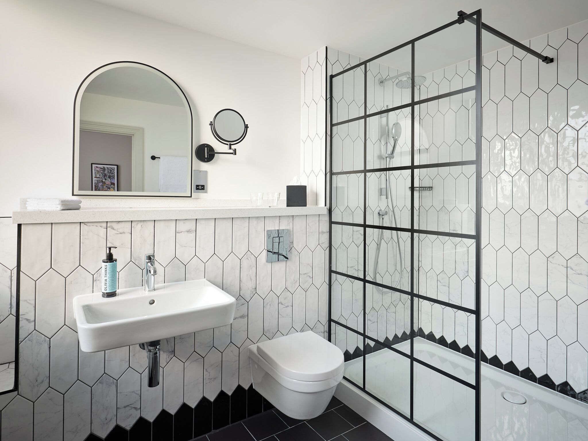 Une salle de bain avec un lavabo blanc, des toilettes, une douche avec des portes en verre et un miroir doté d’un cadre rond.