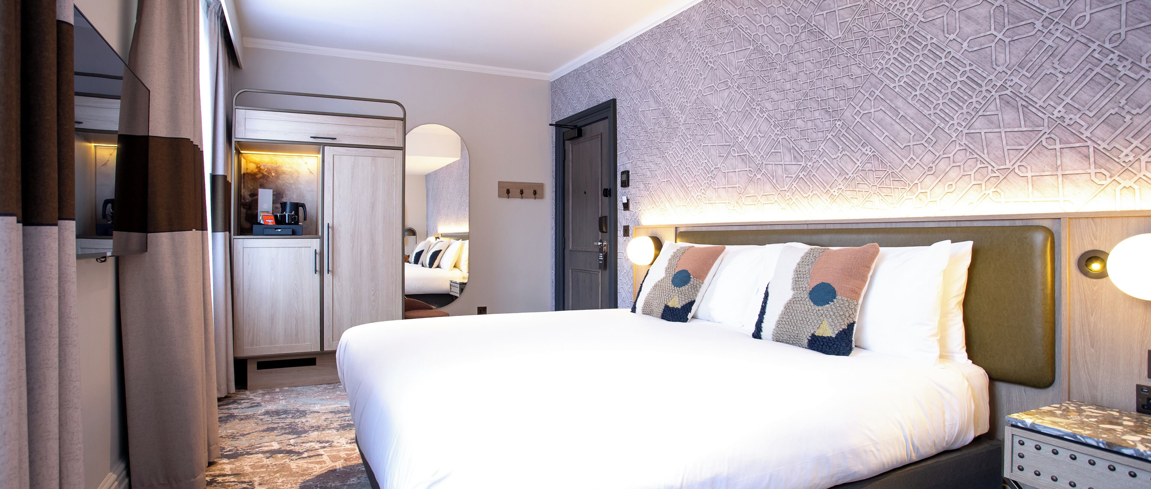 A hotel room with a large bed, a wardrobe, a desk with a coffee maker, and a patterned wall.