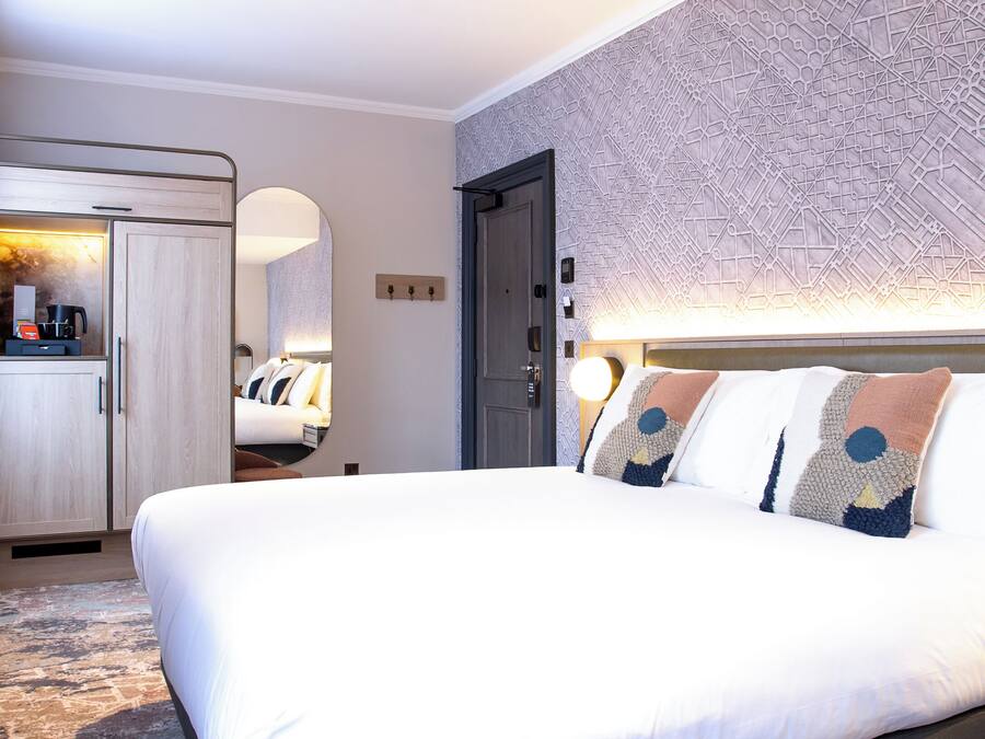 A hotel room with a large bed, a wardrobe, a desk with a coffee maker, and a patterned wall.