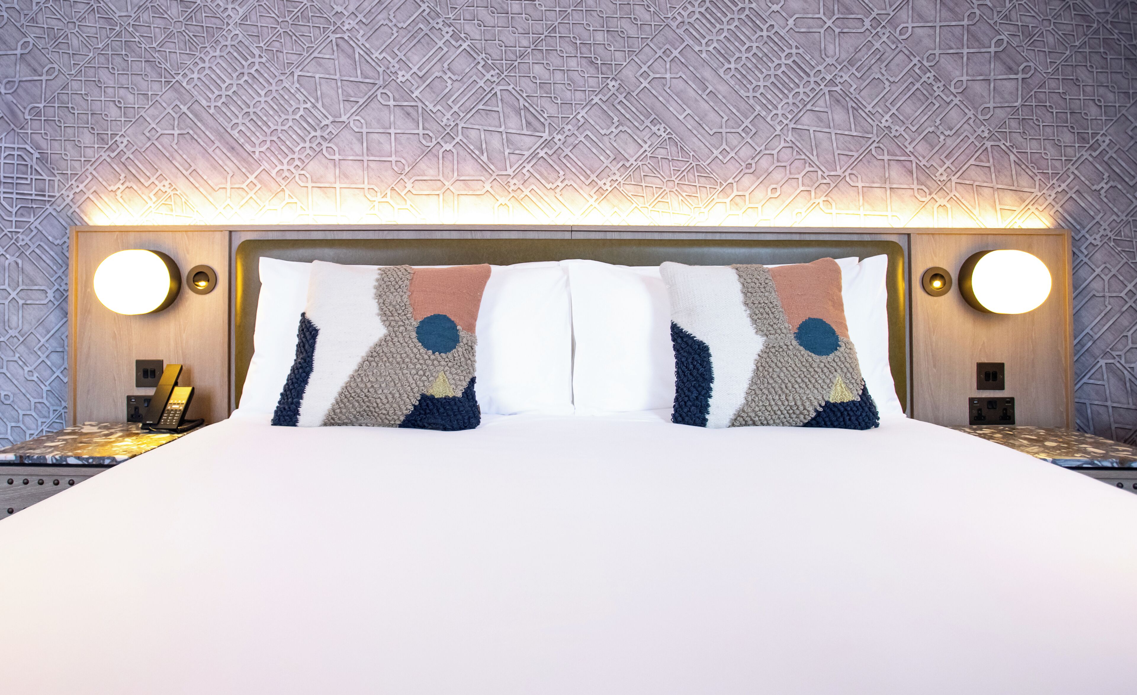 A neatly made bed with a modern headboard, two bedside tables with lamps, and a patterned wall.