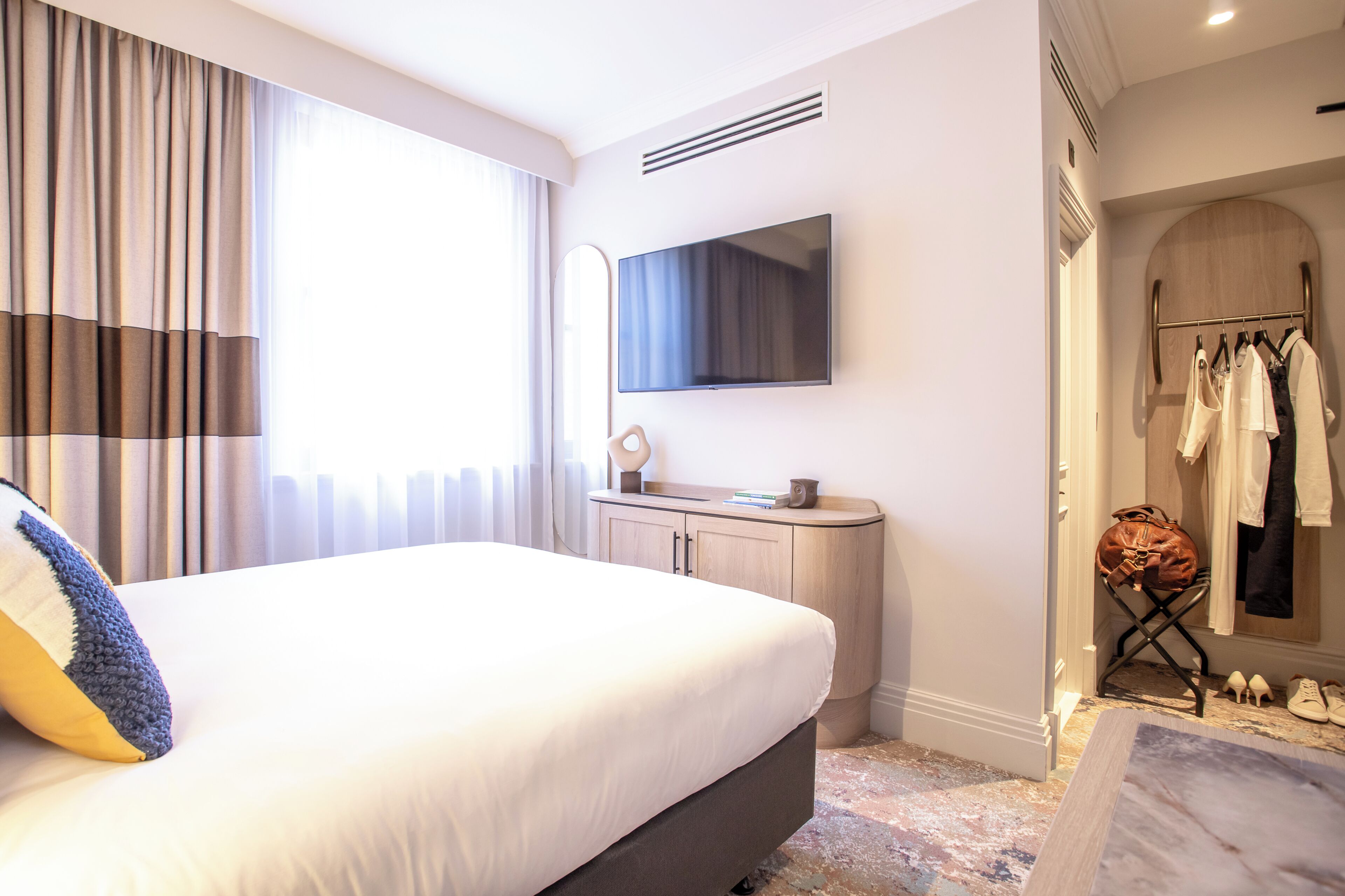A hotel room with a large bed, a flat-screen TV mounted on the wall, a vanity with a mirror, and a walk-in closet with hanging clothes and shoes.