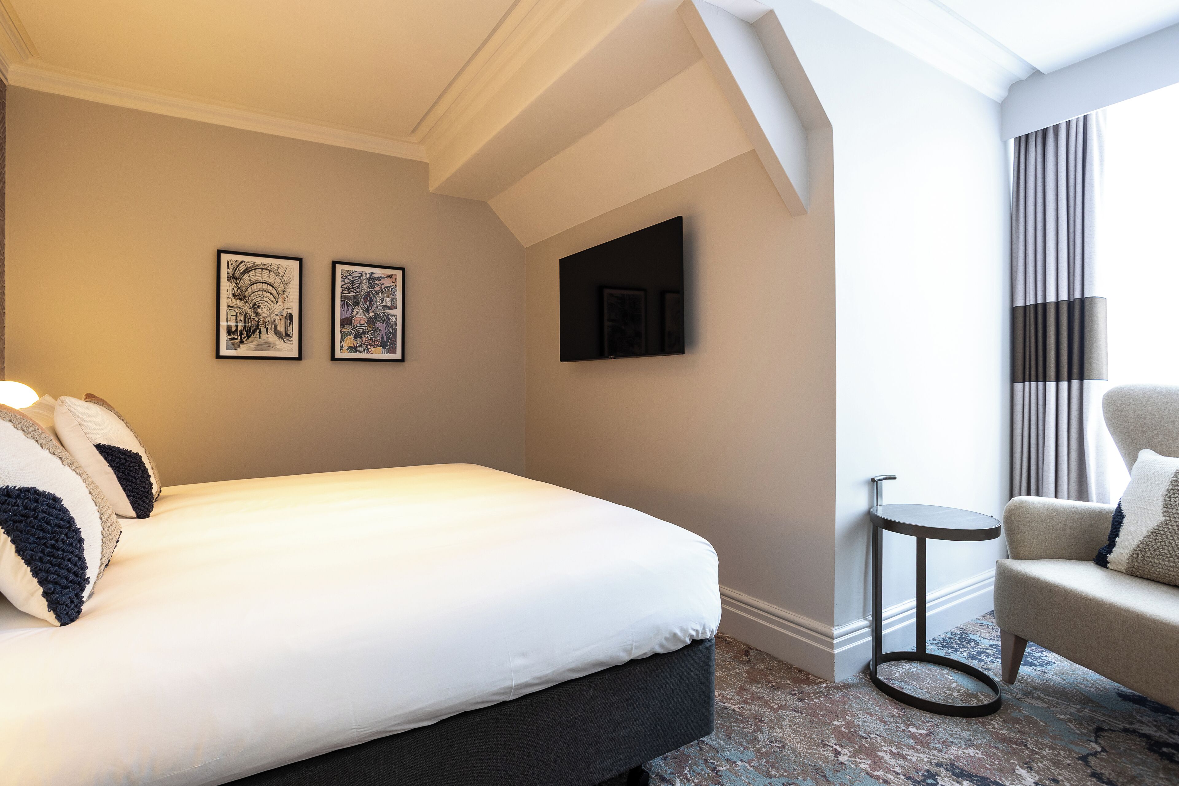 A hotel room with a bed, two framed artworks, a television, a small round table, and a chair.