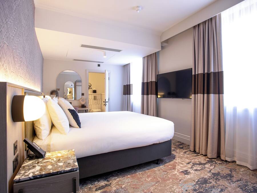 Una moderna camera d'albergo con un letto grande, un comodino con una lampada, una TV a schermo piatto fissata al muro e un'ampia finestra con tende.