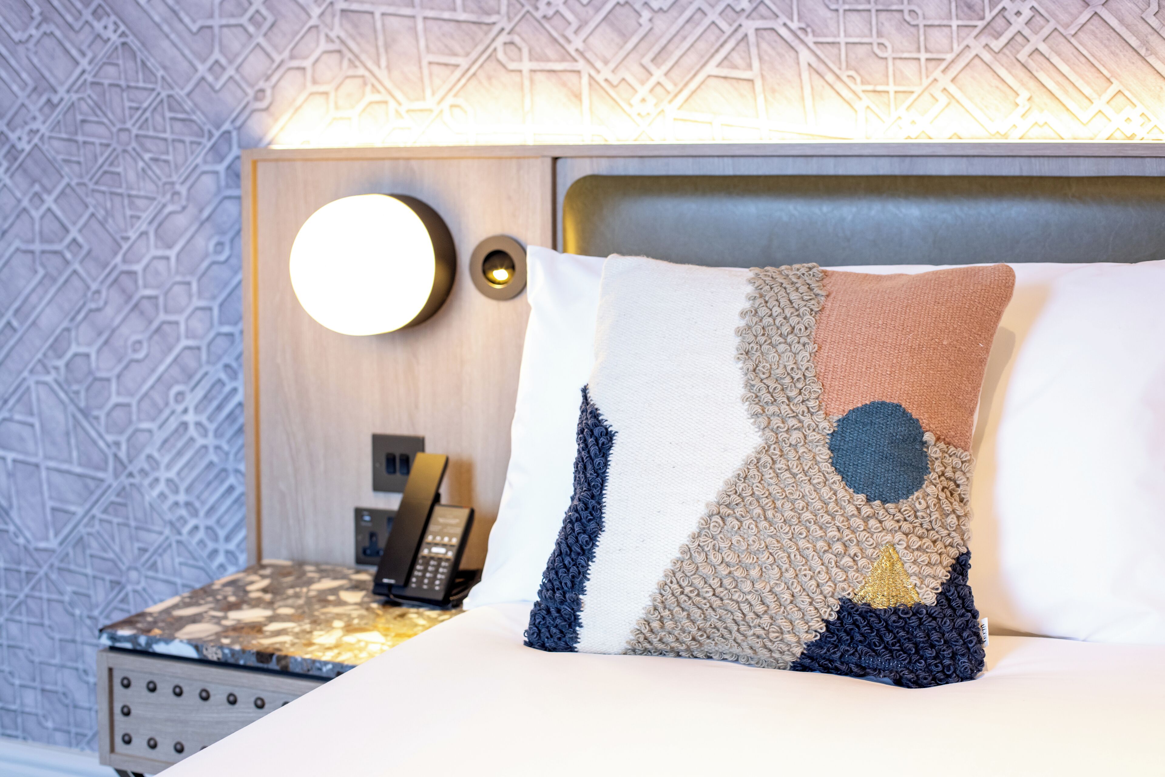 A neatly made bed with a decorative pillow, a bedside table with a phone, and a wall with a geometric pattern.