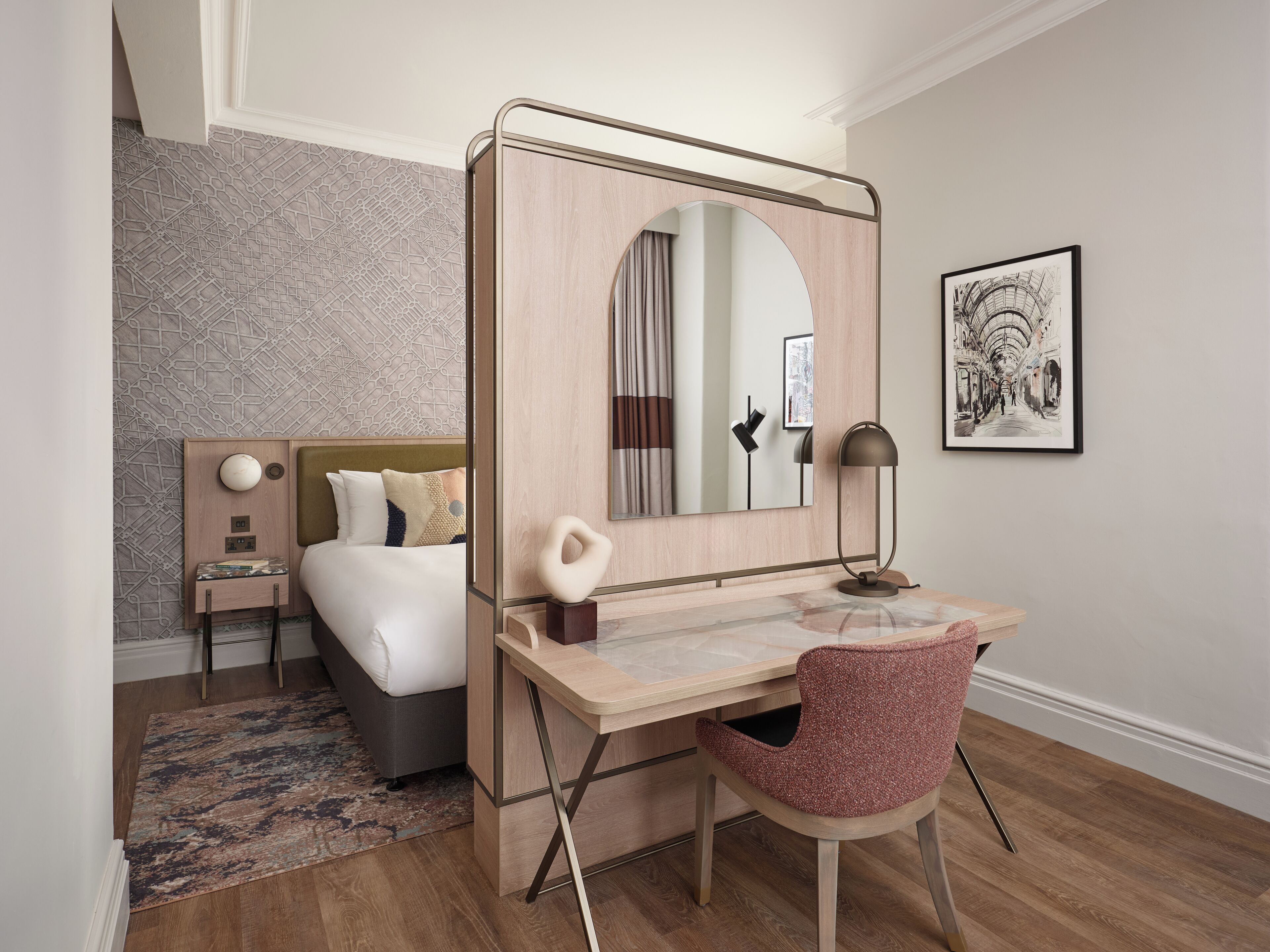 Ein Hotelzimmer mit Bett, Schreibtisch mit Stuhl, Spiegel, Lampe und einem gerahmten Bild an der Wand.