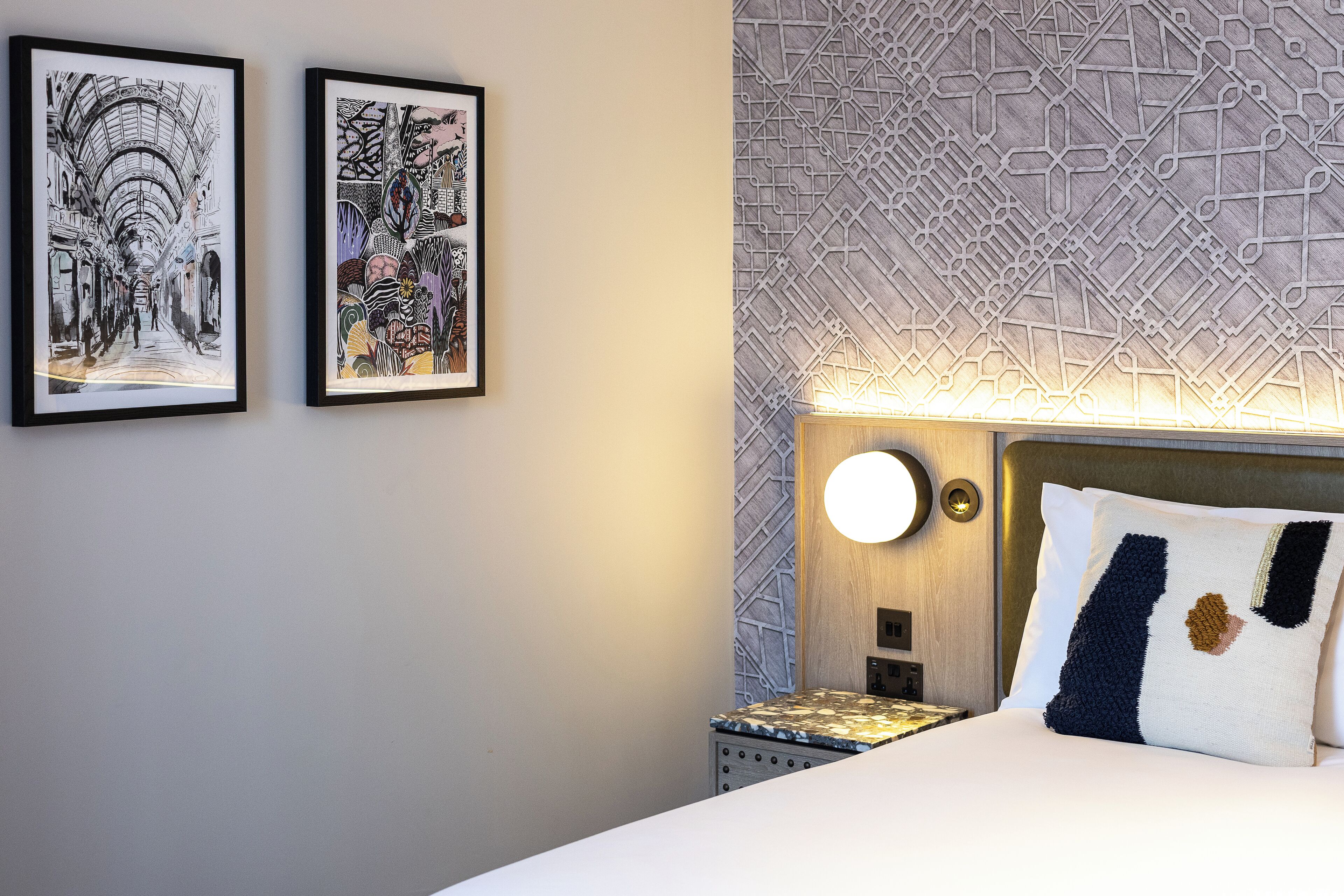 Une chambre d’hôtel comprenant un lit, une table de chevet, une lampe fixée au mur et deux œuvres d’art encadrées accrochées au mur.