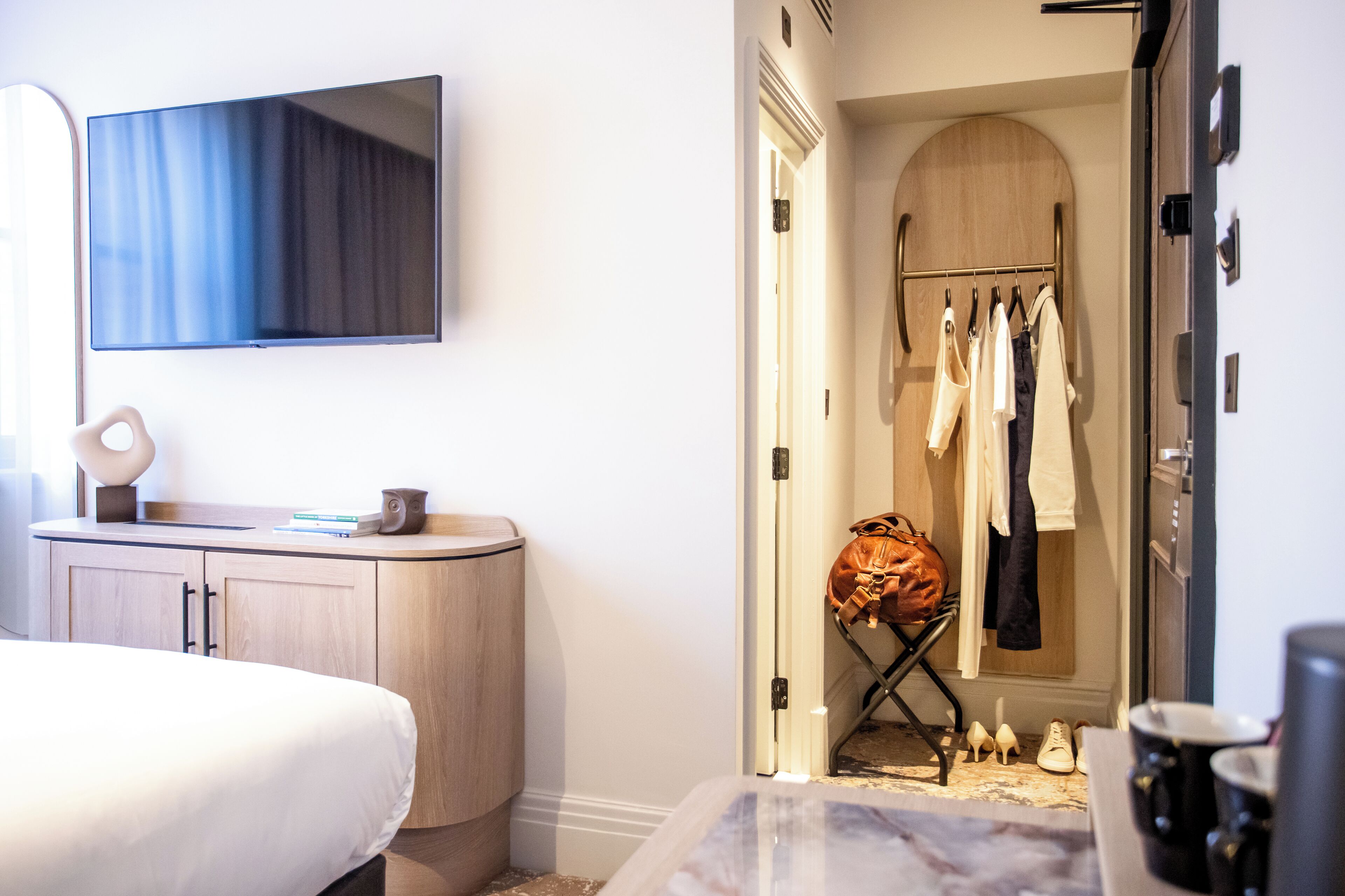 A hotel room with a bed, a TV mounted on the wall, a vanity with a mirror, and a walk-in closet with clothes and shoes.