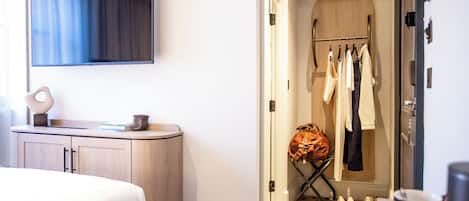 A hotel room with a bed, a TV mounted on the wall, a vanity with a mirror, and a walk-in closet with clothes and shoes.