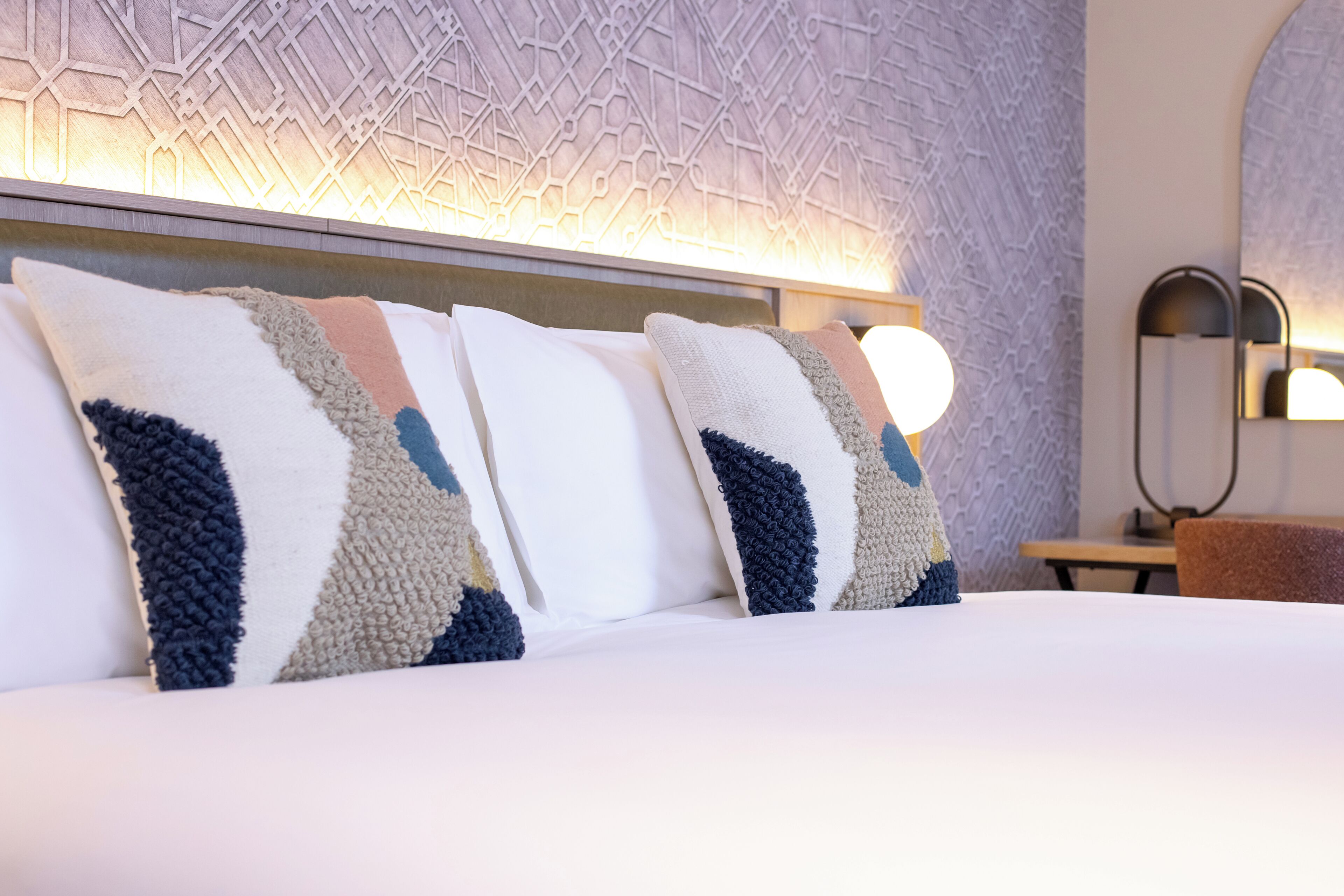 A neatly made bed with patterned pillows, a headboard with a geometric design, and a wall-mounted light fixture.