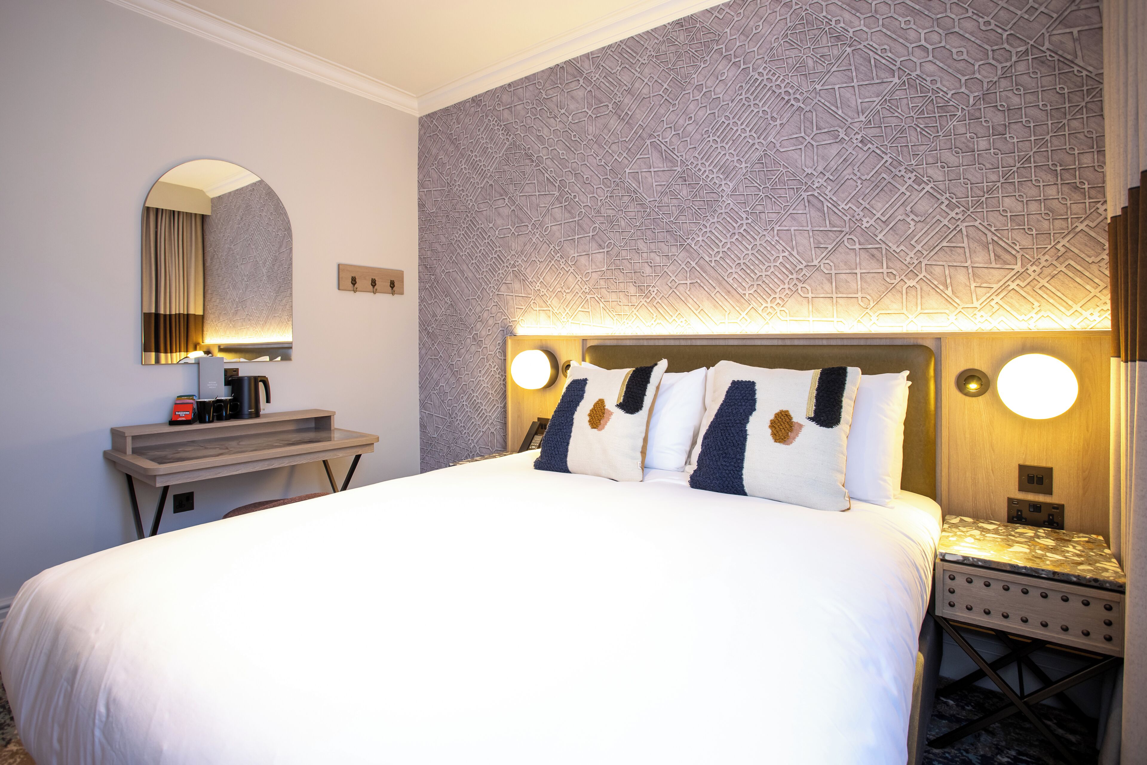 Ein Hotelzimmer mit einem großen Bett, einem Nachttisch, einem Spiegel und einer Wand mit geometrischem Muster.