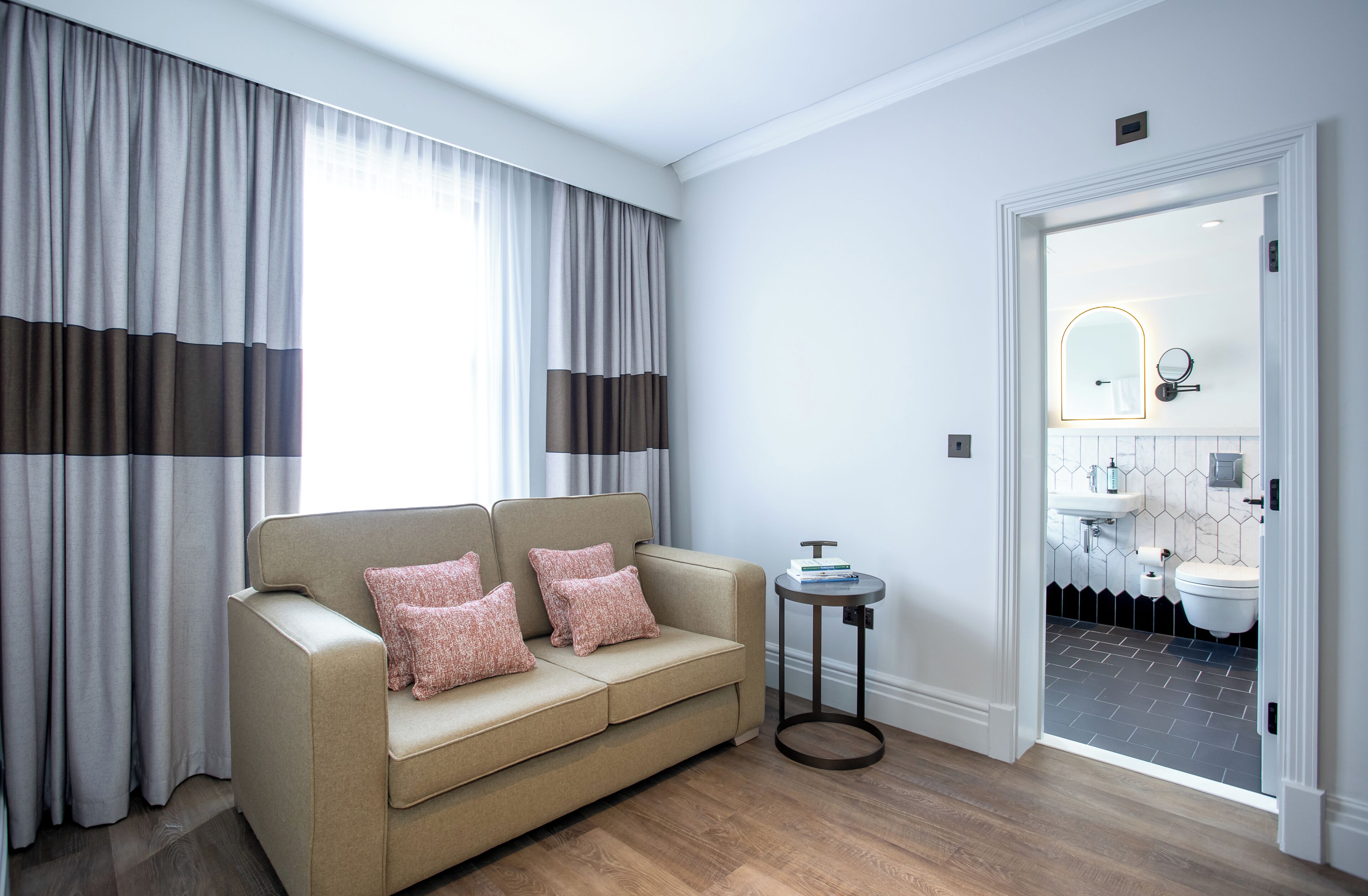 A modern room with a beige sofa, a small round table, and a view of a bathroom through an open door.