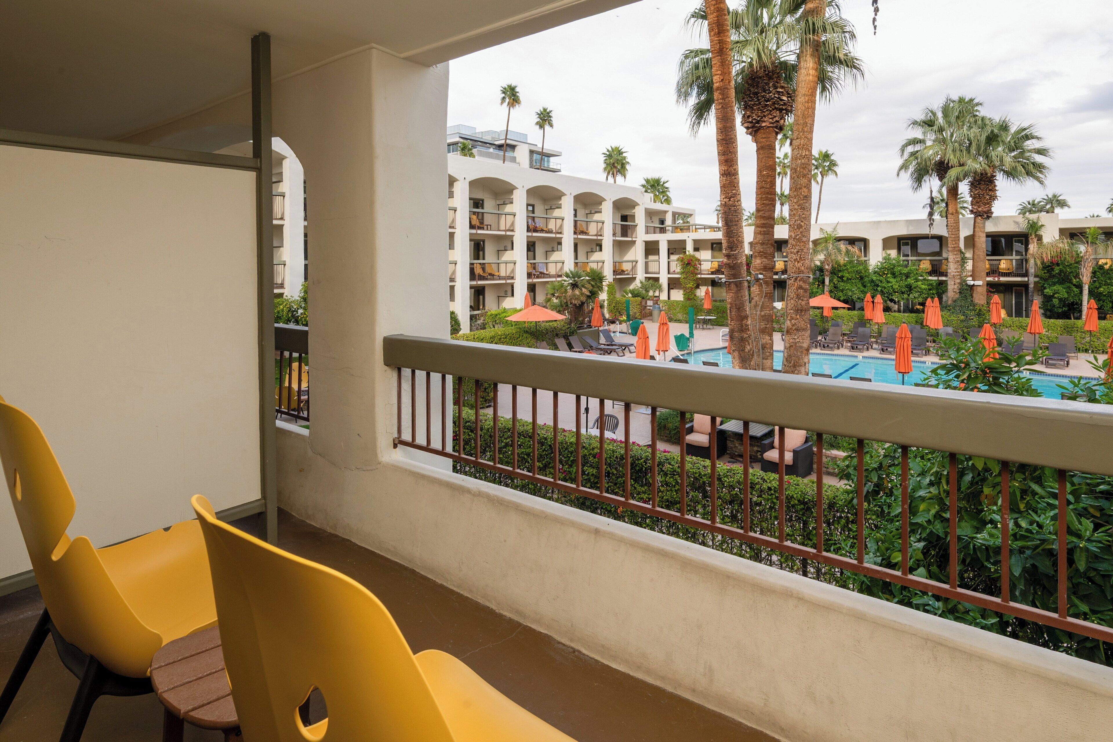 Un balcón de hotel con sillas amarillas, una piscina con sombrillas naranjas y vistas a un edificio de varios pisos con balcones y palmeras.