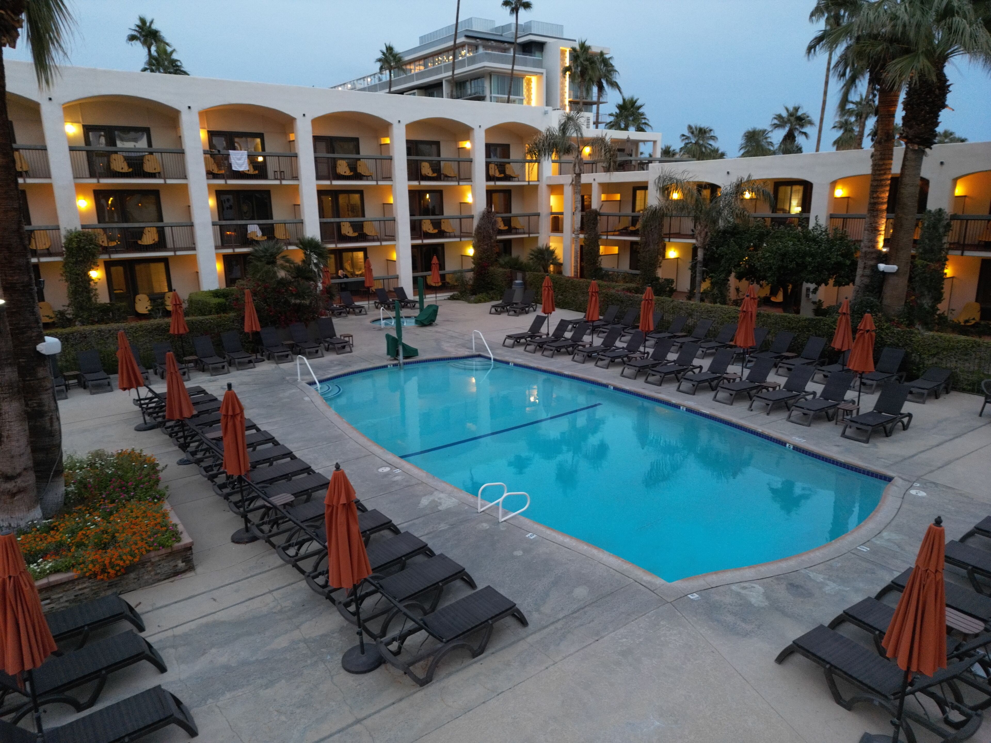 A hotel pool area with lounge chairs and umbrellas, surrounded by palm trees and a building with multiple balconies.