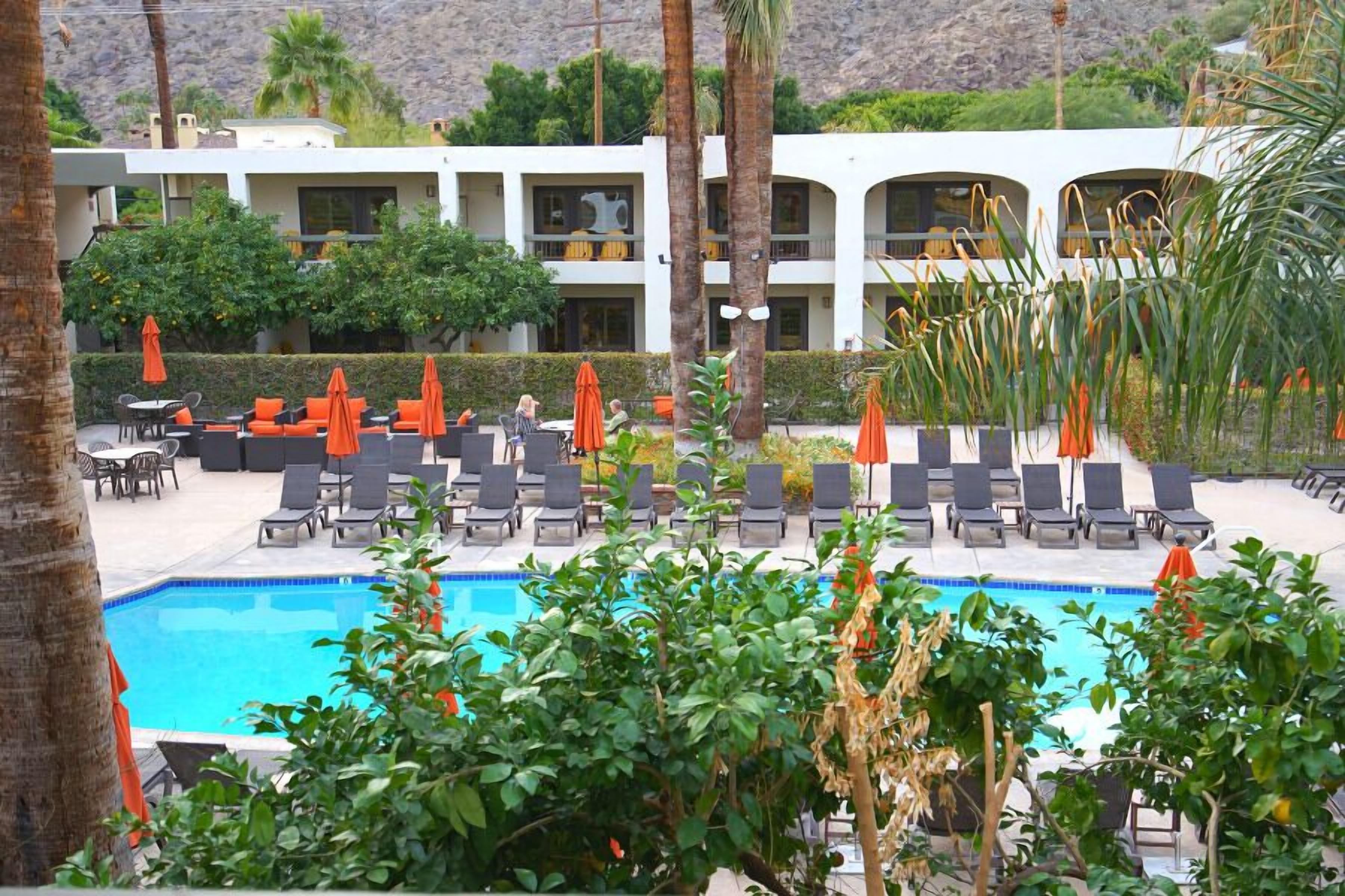 A pool area with lounge chairs and umbrellas, a building with multiple balconies, and a mountainous backdrop.