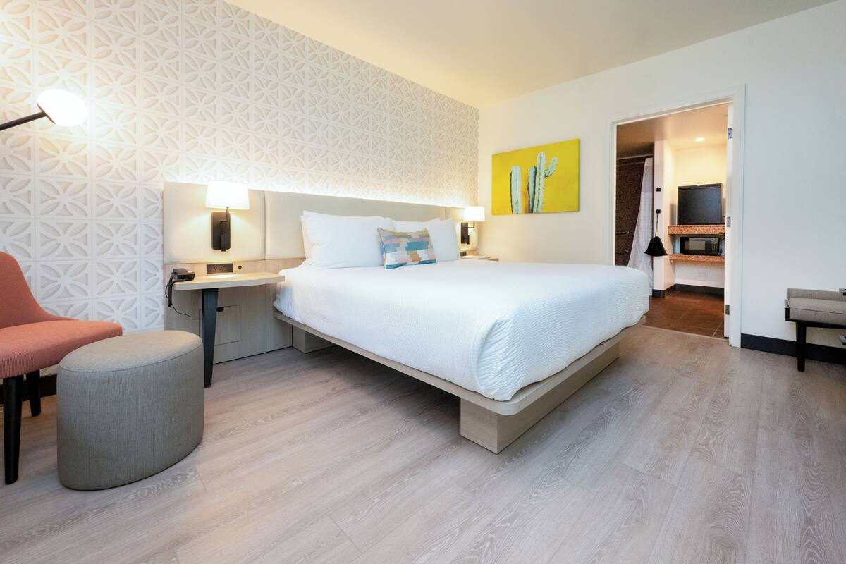 A modern hotel room with a large bed, bedside tables, a chair, and a small ottoman.