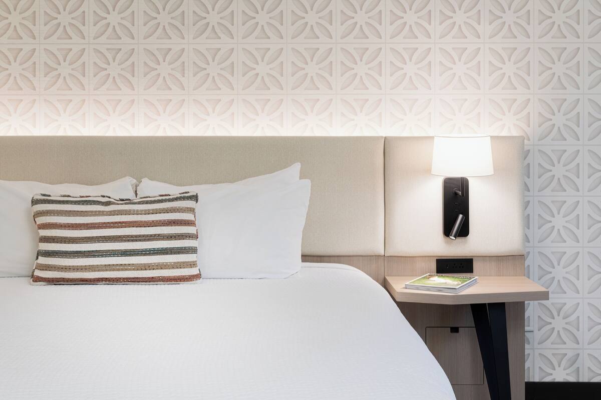 A hotel room with a neatly made bed, a patterned pillow, a bedside table with a lamp, and a decorative wall panel.