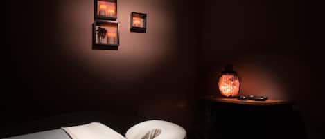 Una habitación tenuemente iluminada con una cama, un almohadón blanco y una mesita de noche con una lámpara.