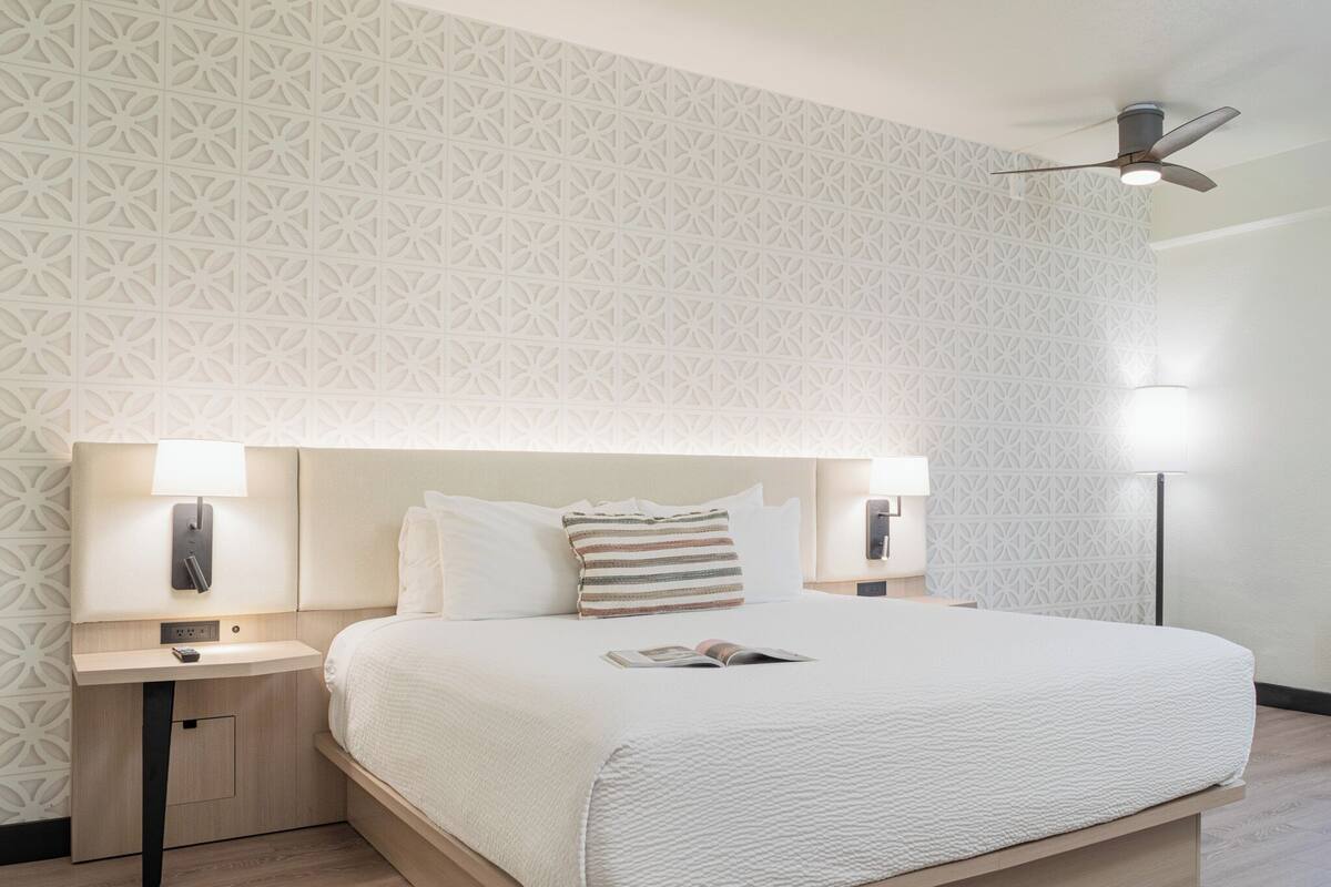 A modern hotel room with a large bed, two bedside tables with lamps, a ceiling fan, and a patterned wall.
