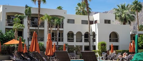Zona de piscina con sillones de descanso y sombrillas, palmeras y un edificio con balcones.