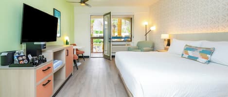 Habitación de hotel con cama, televisión, escritorio con computadora, silla y ventana con vista a palmeras.