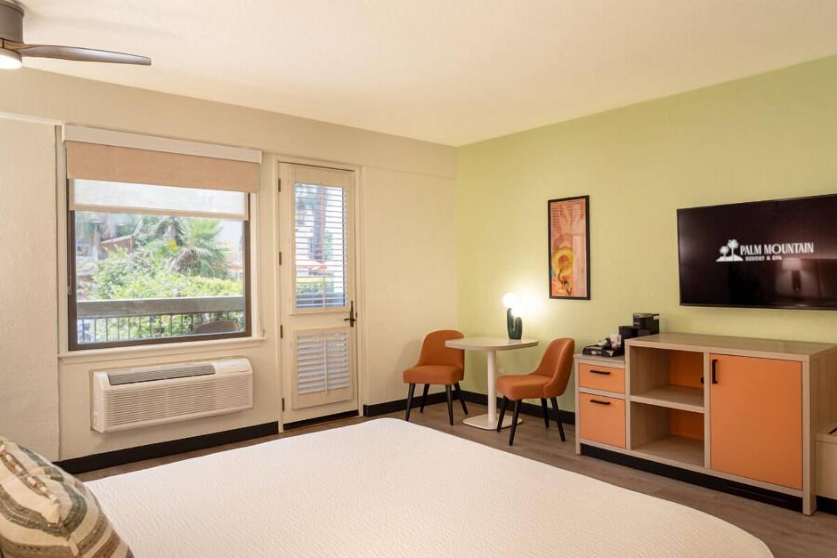A hotel room with a bed, a desk with two chairs, a television, and a large window with blinds.