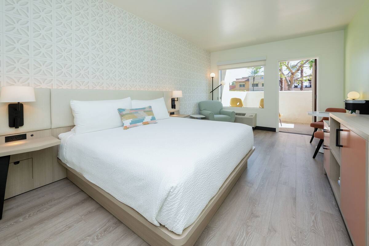 A modern hotel room with a large bed, a desk, a chair, and a view of the outside.